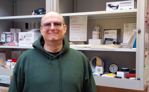Philip smiling, wearing a green sweatshirt and glasses, standing in front of an open cabinet with stationary and assistive technology.