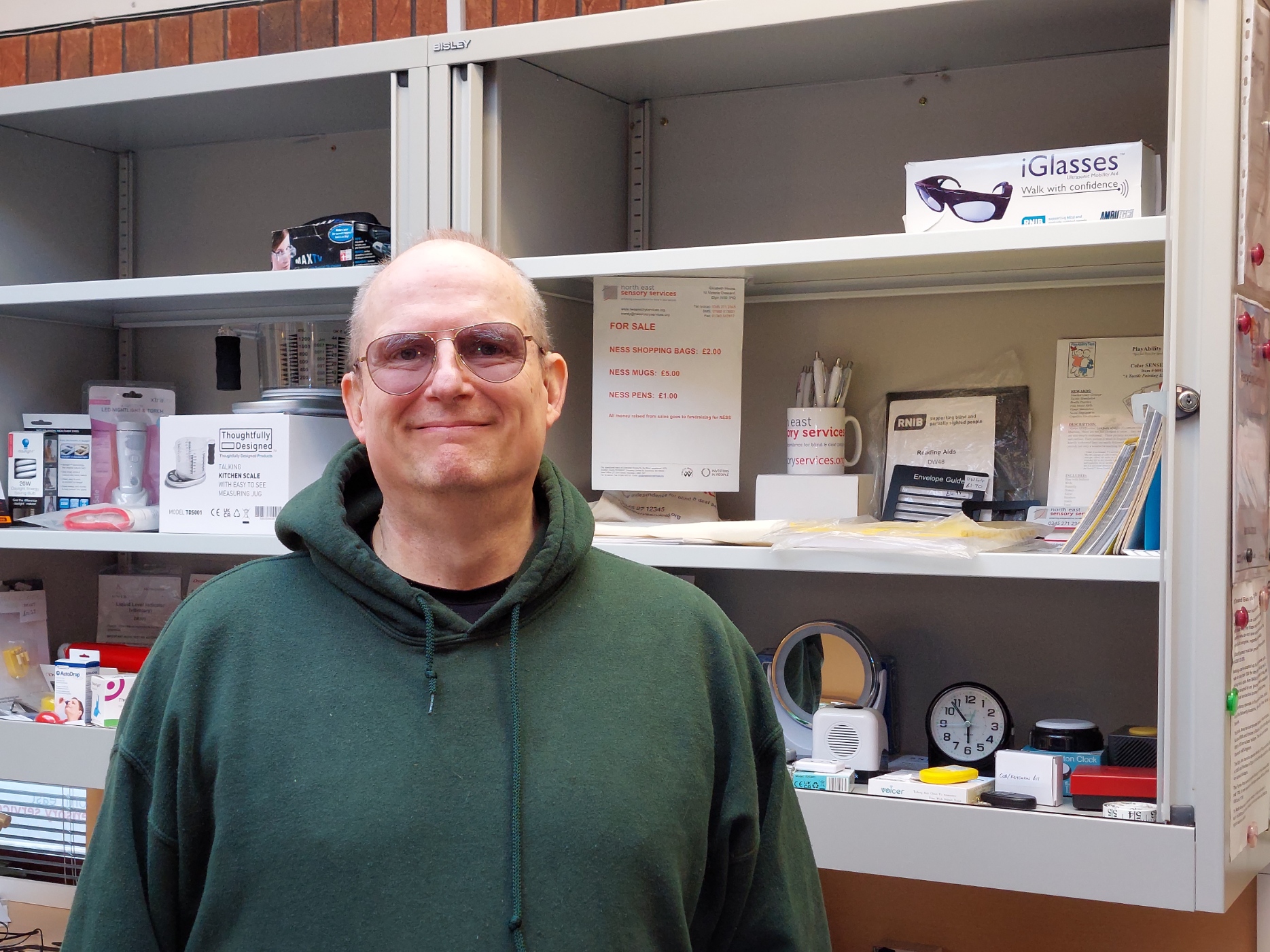Philip smiling, wearing a green sweatshirt and glasses, standing in front of an open cabinet with stationary and assistive technology.