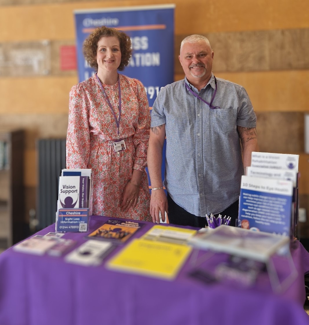 Our Sight Loss Information Officers, Ash and Hayley smiling and standing in front of a table full of information leaflets.