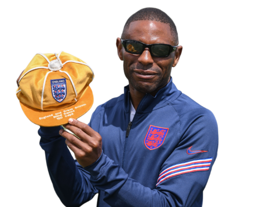 Darren wearing his navy England tracksuit and holding a gold cap with the three lions crest, and the words England and Great Britain blind squad, 100 caps. Darren wearing his navy England tracksuit and holding a gold cap with the three lions crest, and the words England and Great Britain blind squad, 100 caps.