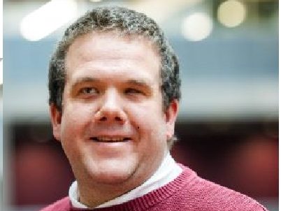 Robbie, a white male with short hair, stands in the foreground with a blurry office-type scene in the background. Robbie is smiling slightly at the camera and wearing a maroon jumper with a white collar peeking through. Only his head and shoulders are in view. Robbie, a white male with short hair, stands in the foreground with a blurry office-type scene in the background. Robbie is smiling slightly at the camera and wearing a maroon jumper with a white collar peeking through. Only his head and shoulders are in view.