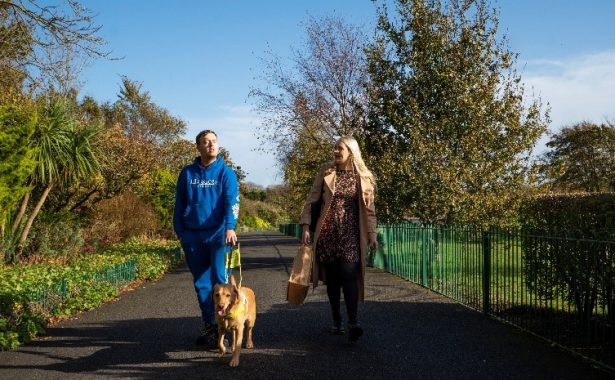A vision impaired person with their guide dog and a member of the Vision Support Barrow & District team walking side by side through Barrow Park. Image Credit: Vision Support Barrow & District / Phil Green