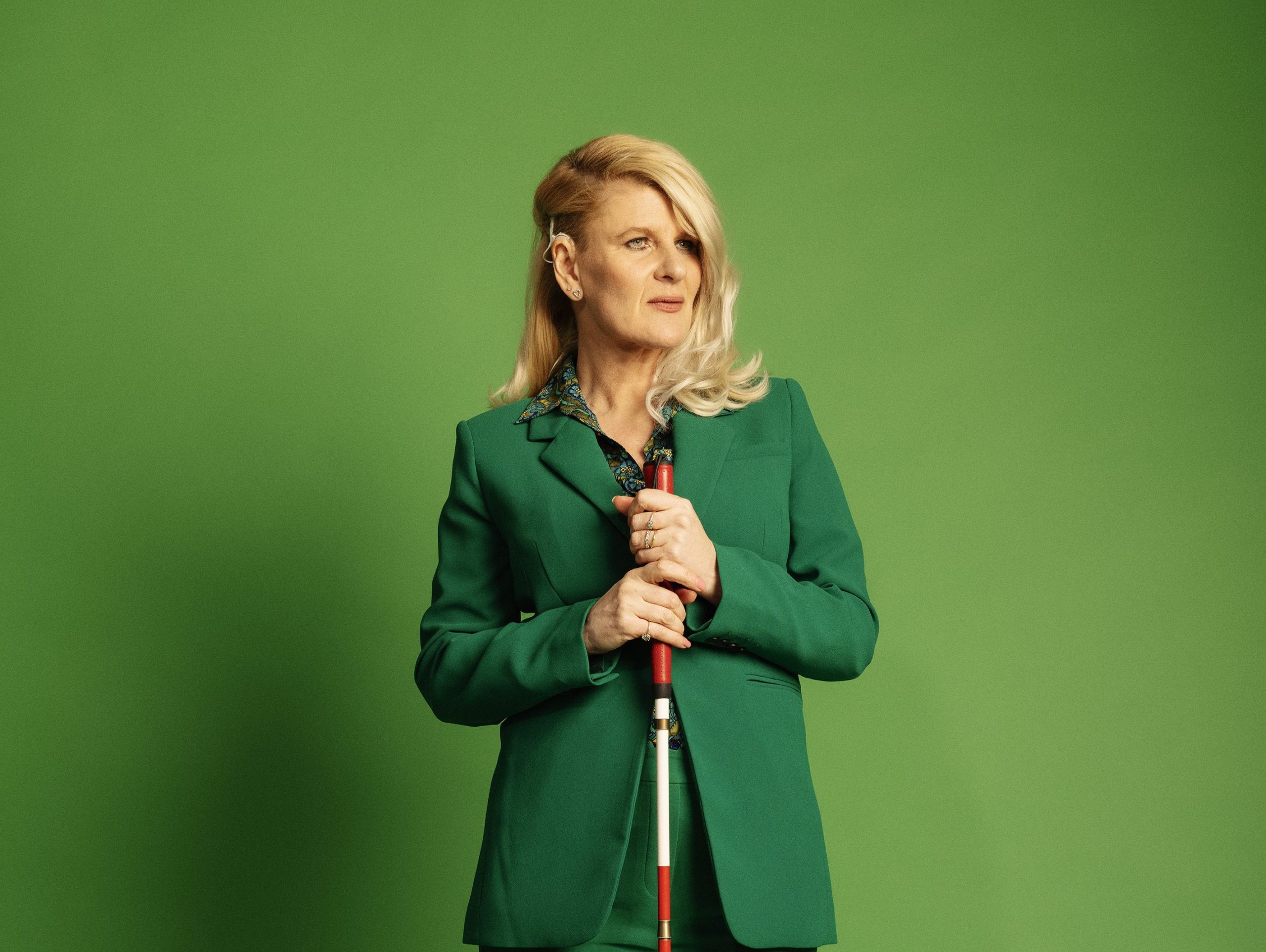 Jo is standing holding her red and white striped mobility cane. Jo is wearing a green trouser suit and has blonde mid length hair. A cropped version of a full length photo of Jo holding her red and white striped mobility cane. Jo is wearing a green trouser suit and has blonde mid length hair,