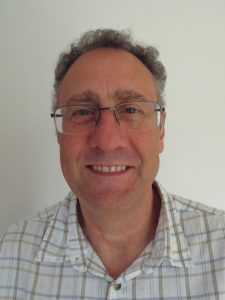 Carl Hodge. He is smiling towards camera and wears a checked shirt and glasses.