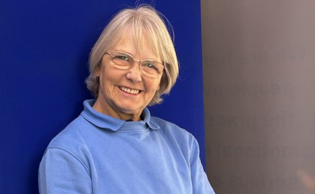 Ethna is smiling, standing against a blue background. She has short blonde hair, is wearing glasses, a blue top and jeans.