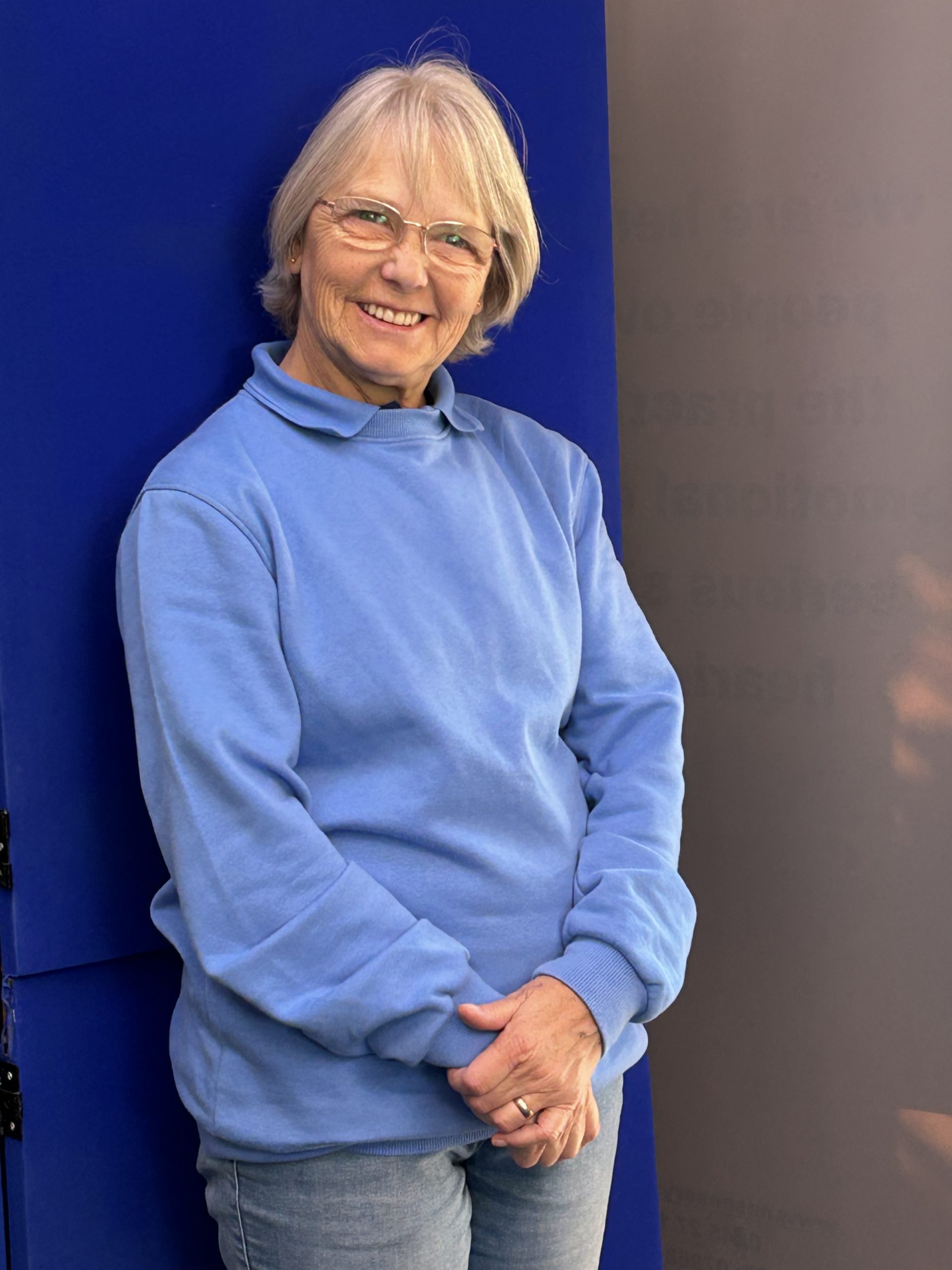 Ethna is smiling, standing against a blue background. She has short blonde hair, is wearing glasses, a blue top and jeans.