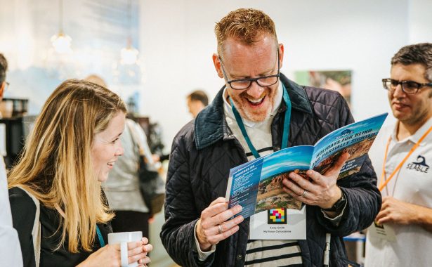 Andy and Joanna, from MyVision Oxfordshire, smiling whilst reading the Seable brochure. Daniel from Seable in on the right of picture, behind Andy. There are other delegates and exhibitors in the image either blurred or with their backs to the camera.