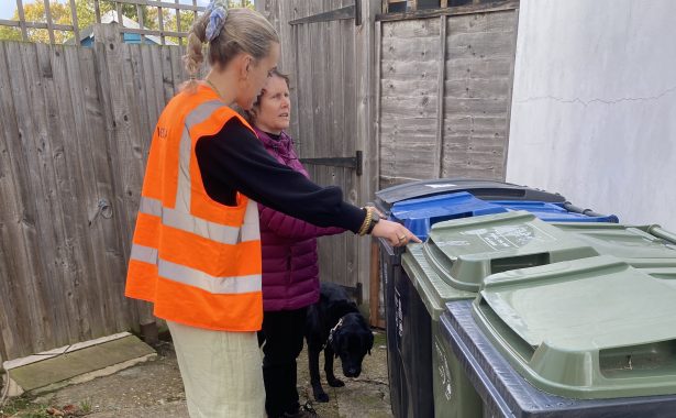 A lady in a high vis vest with a lady who has a guide dog in a garden with a row of bins.