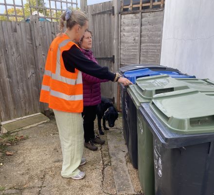 A lady in a high vis vest with a lady who has a guide dog in a garden with a row of bins.