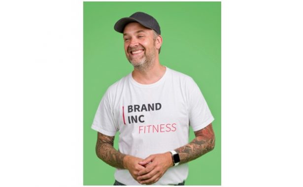 Richard Pinchen has a large, warm smile. He is wearing a baseball cap, watch and white T-shirt with the Brand Inc Fitness logo on it. Richard has a beard and tattoos on his arms.