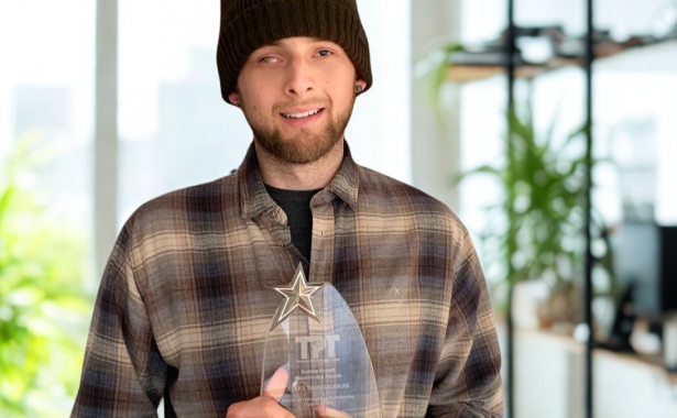 Aleks wears a brown checked shirt and a beanie, and smiles to the camera whilst holding a glass award.