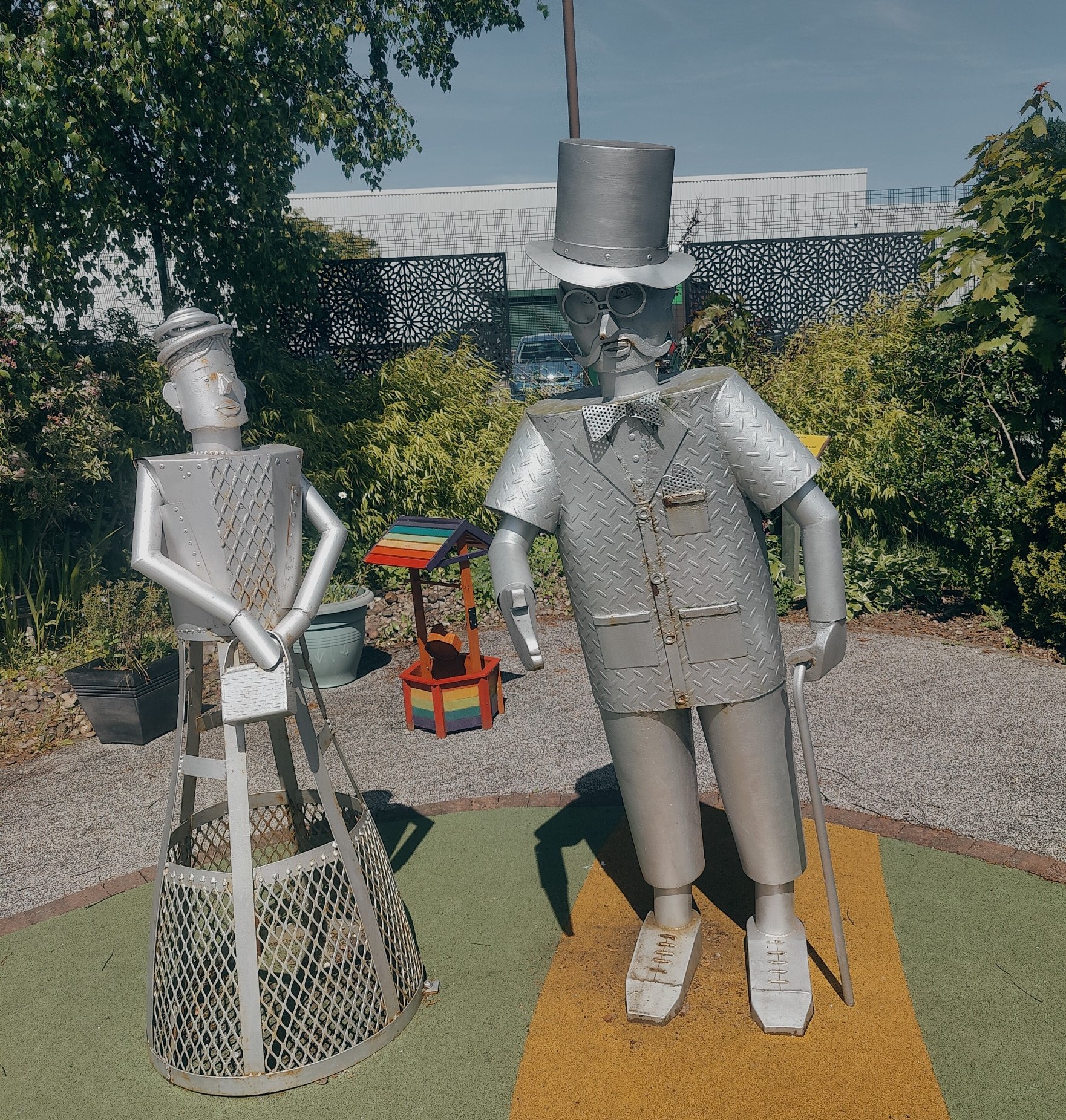 A full-length picture of Douglas the Tin Man with the Tin Lady, known as Cathie, in the sensory garden on a sunny day.