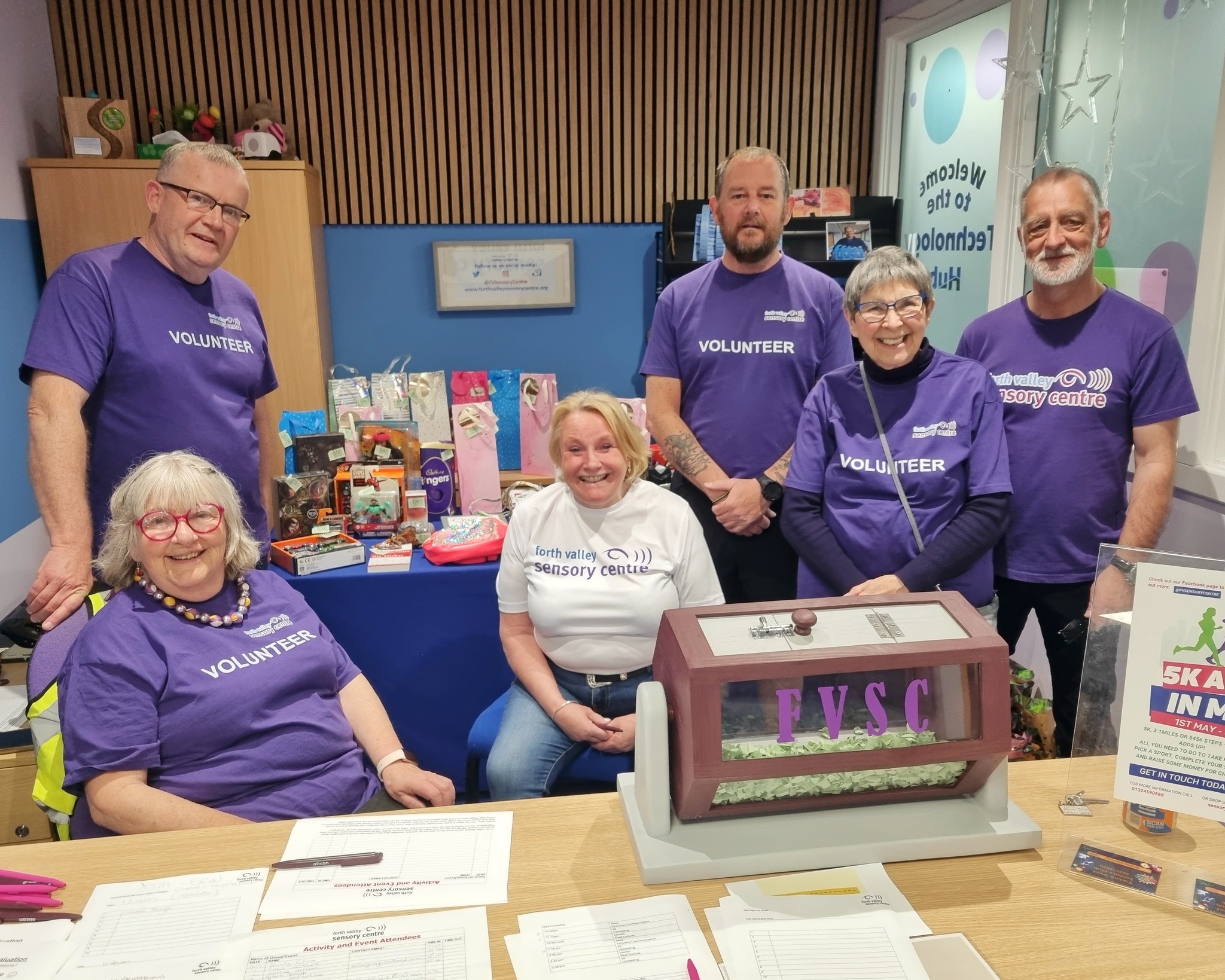 A group photograph shows (left to right) volunteers behind a desk who are wearing Forth Valley Sensory Centre T-shirts: John Somerville, Christine Fraser, Jenn Pow, Duncan McPhee, Ann McFarlane, and Colin Watterson.