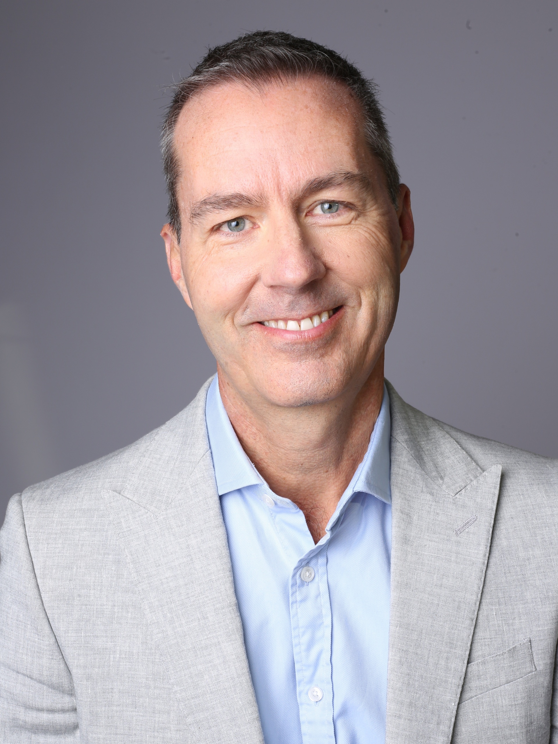 Richard smiles warmly with head slightly tilted to the side. He is wearing a light blue shirt and light grey blazer.