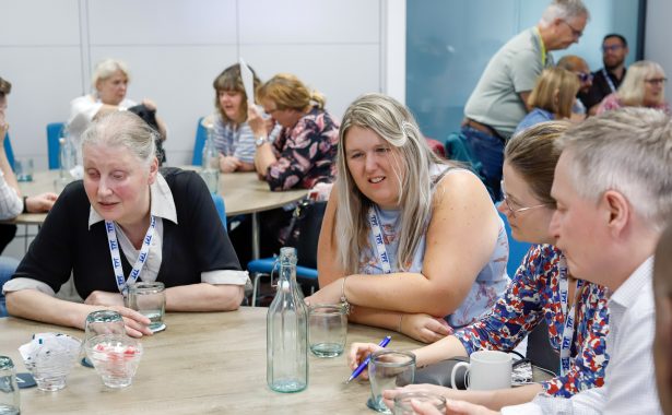 Mary, Leanne, Vicky, and Harry, Sight Loss Council volunteers, sit around a table at the annual SLC Conference taking part in a roundtable discussion, while other volunteers are seated at nearby tables in the background doing the same.