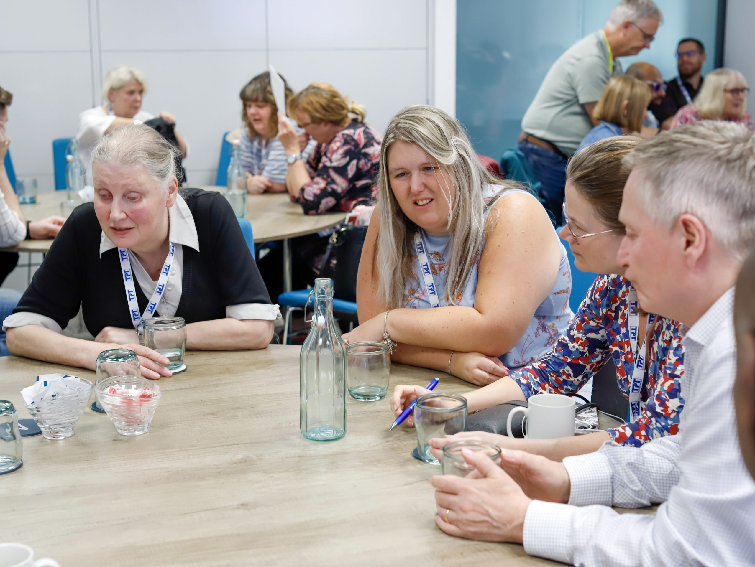 Mary, Leanne, Vicky, and Harry, Sight Loss Council volunteers, sit around a table at the annual SLC Conference taking part in a roundtable discussion, while other volunteers are seated at nearby tables in the background doing the same.