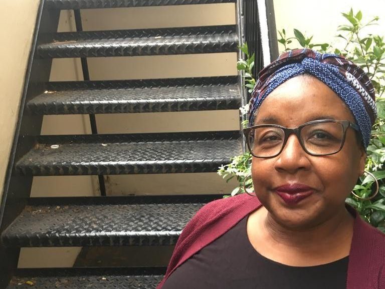 Beverley is smiling. She is a black woman wearing a blue hair band, glasses, a black top and burgundy cardigan. There are stairs and a plant in the background.