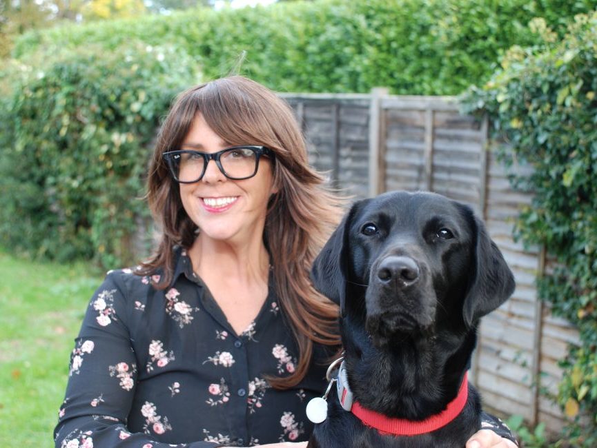 Clare is smiling. She has long, layered brunette hair and a fringe. She is wearing glasses and a black shirt with flowers. Her black dog is by her side wearing a red collar.