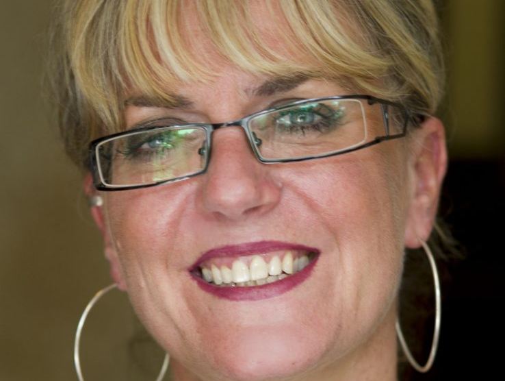 Debra has a blonde fringe, is wearing glasses and large hoop earrings and is smiling.