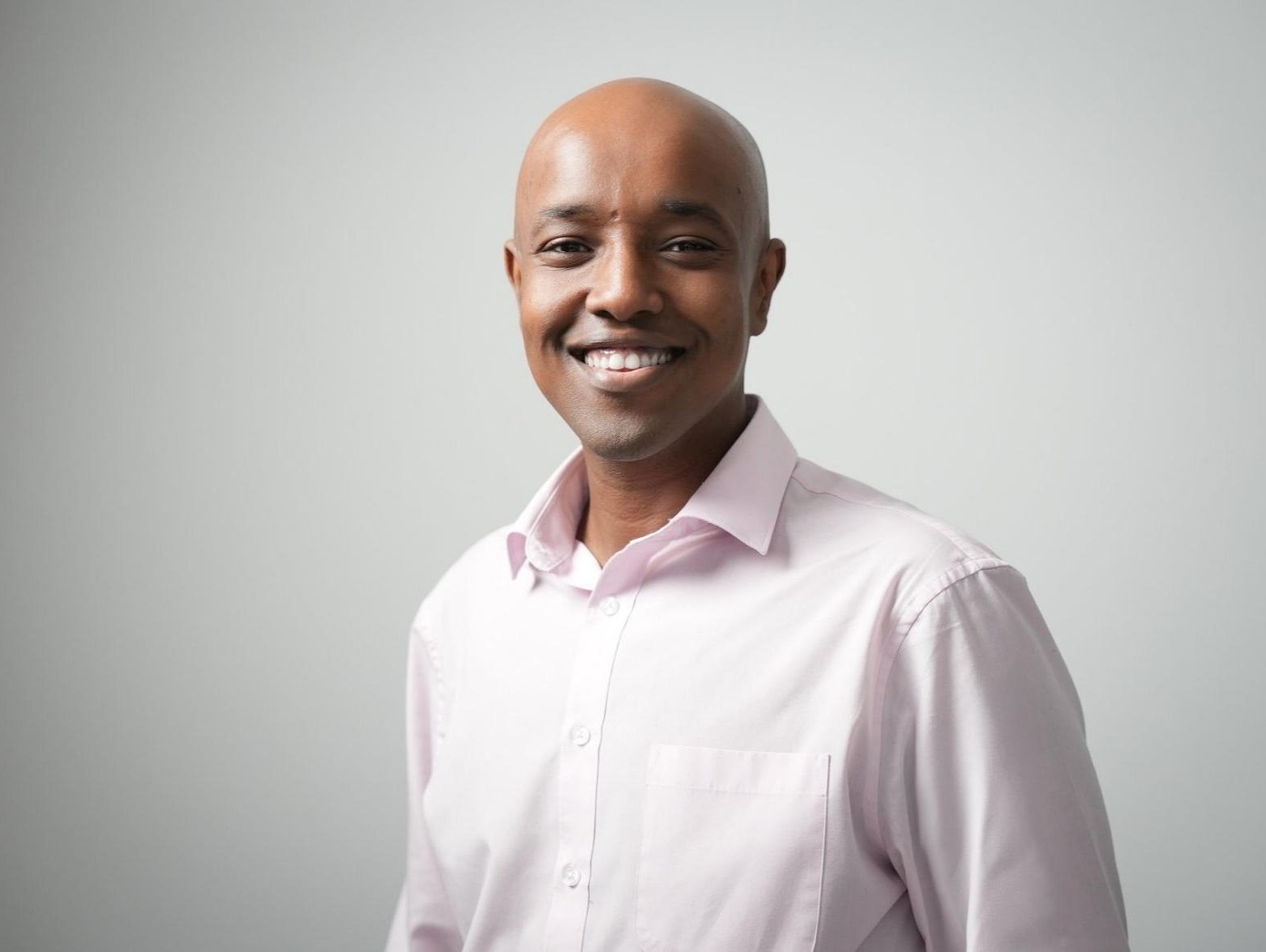 Harbi is a young, black man with a shaved head and a big smile. He is wearing a light pink shirt with the top button open.