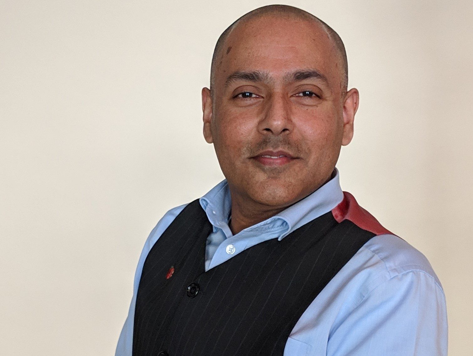 Kamran is a young Asian man with a shaved head and is smiling. He is wearing a light blue shirt and dark blue waistcoat.