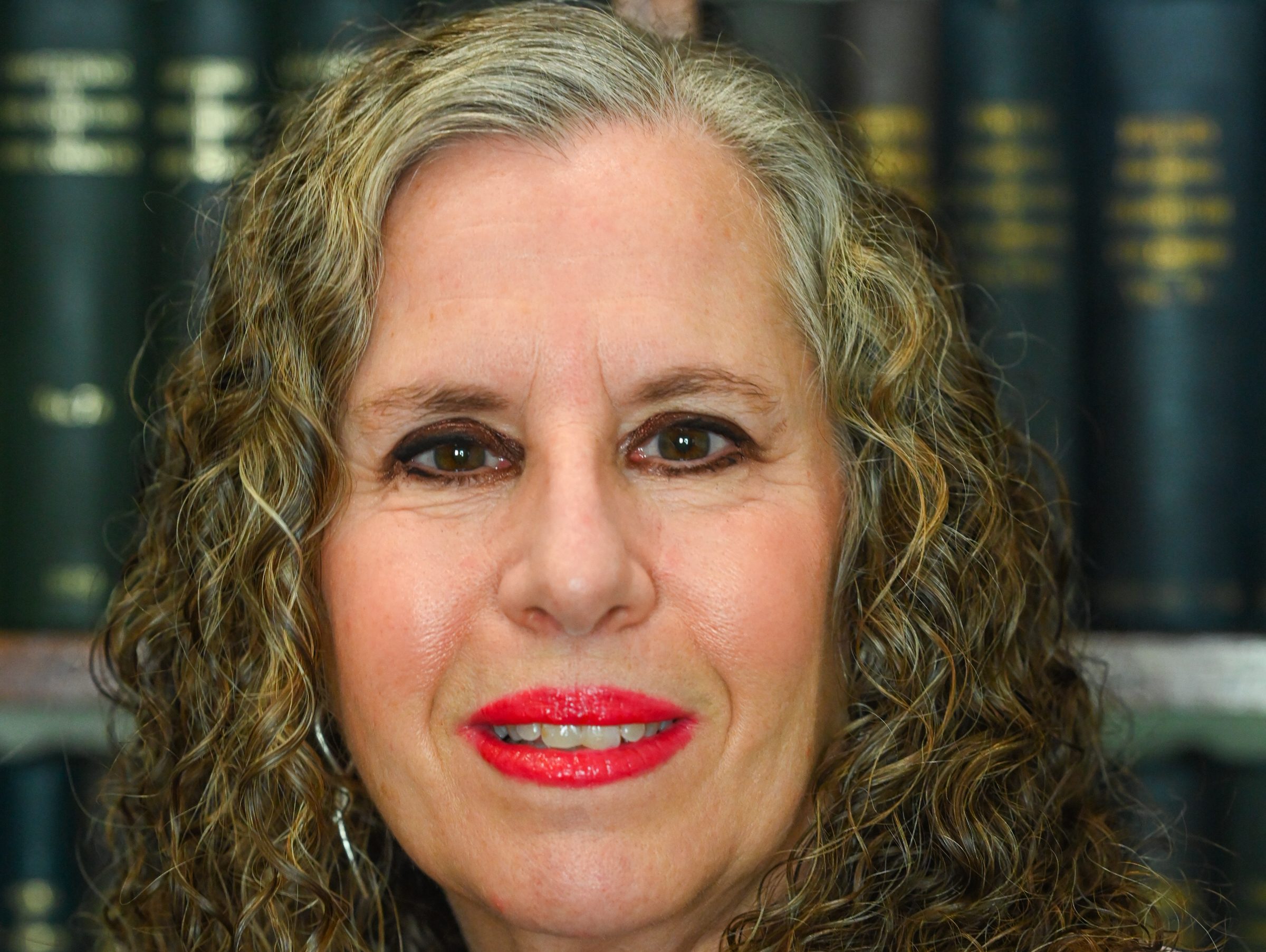 Sandi has curly brown hair and is smiling. She is wearing red lipstick and a patterned black and white top.