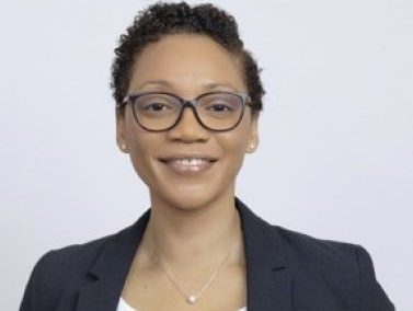 Susanette is a black woman who has short, black hair and is smiling. She is wearing glasses, diamond stud earrings, a gold chain necklace with a pearl, a white top and black blazer.