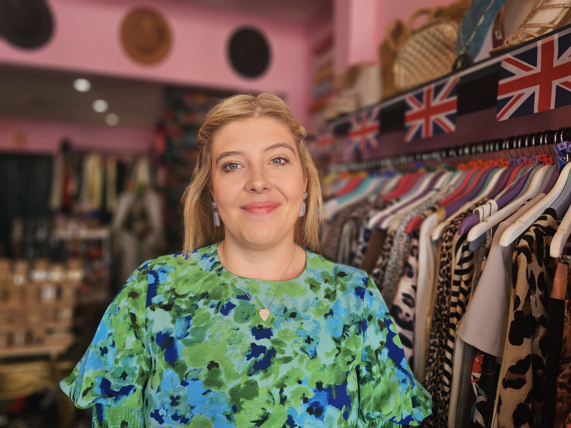 Tilly has long blonde hair pulled back at the sides and is smiling. She is wearing a colourful green and blue top with a heart pendant. She is standing in a shop by a clothes rail and union jack flags above this.
