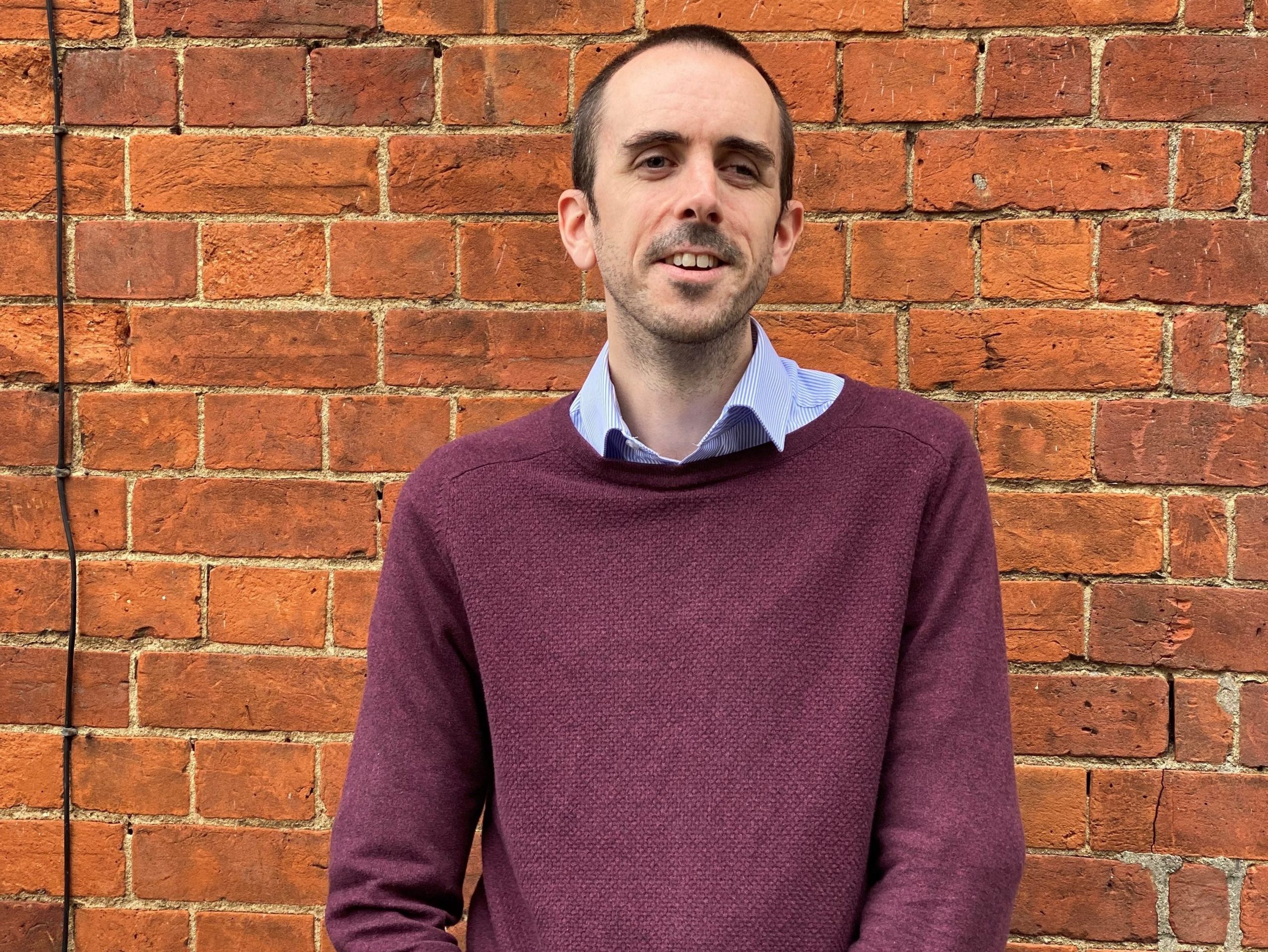 Mark is smiling, standing against a brick wall. He is wearing a burgundy jumper and blue shirt.