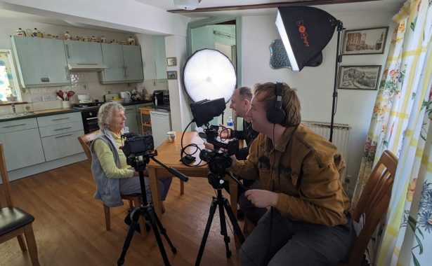 A mature lady wearing yellow and grey sports outfit is sitting at a table in her kitchen, being filmed and interviewed by two men.