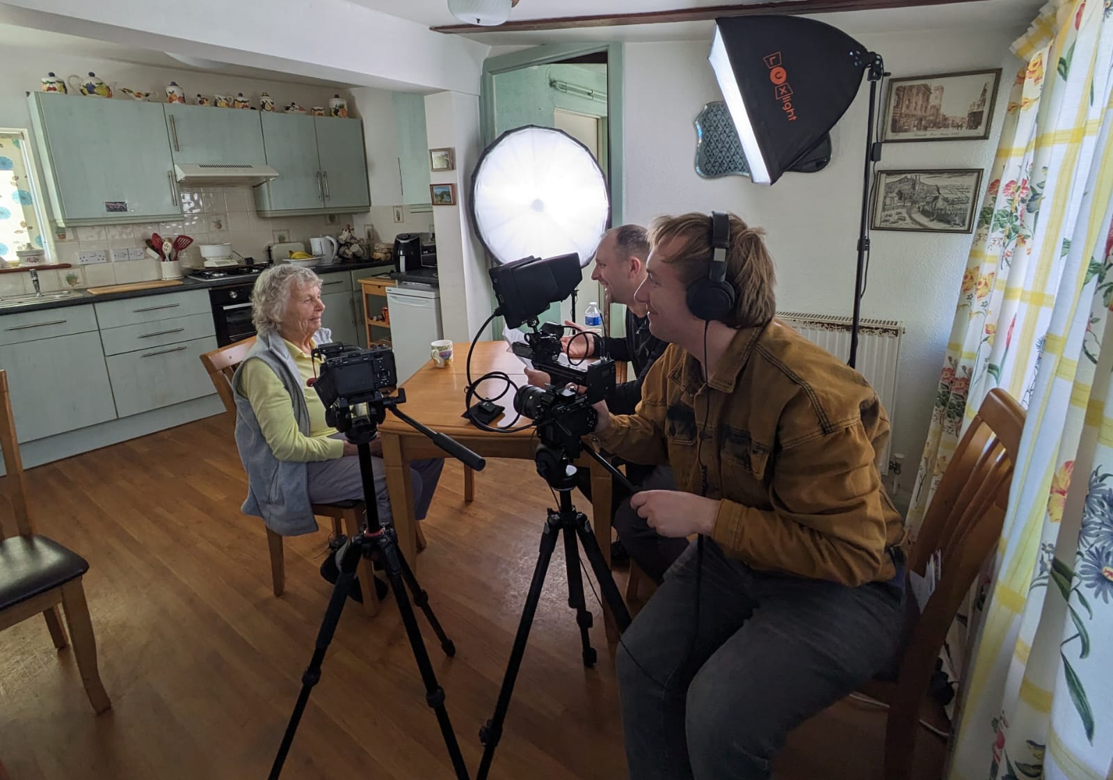 A mature lady wearing yellow and grey sports outfit is sitting at a table in her kitchen, being filmed and interviewed by two men.