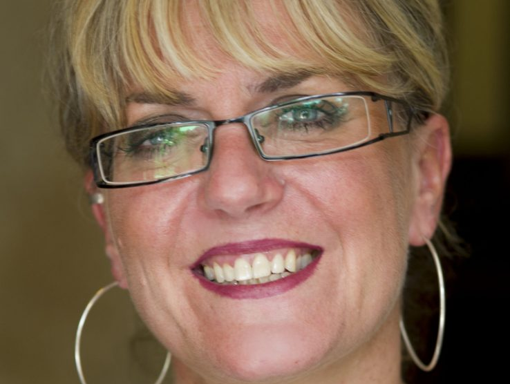 The face of a white woman in her early sixties with white blonde hair wearing big glasses, and large silver hoop earrings, the background is blurred.