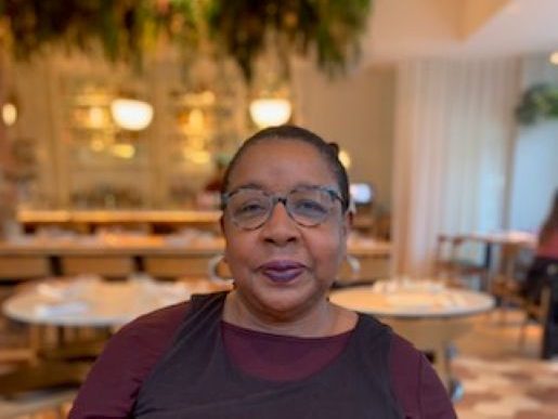 Head and shoulders shot of Black woman (brown skin), smiling, with short black/grey hair. Wearing glasses, a brown long sleeved top with black overtop. The background is a cafe with two white table and warm lighting.
