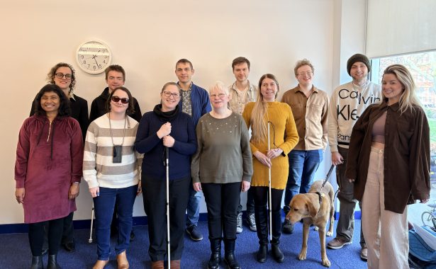 Group shot of 12 male and female interns from the 2025-26 cohort stood at the back of an office room with a blue carpet and cream walls. A guide dog can be seen in the photo. Several of the interns are holding a white navigation cane. They are smiling and look happy.