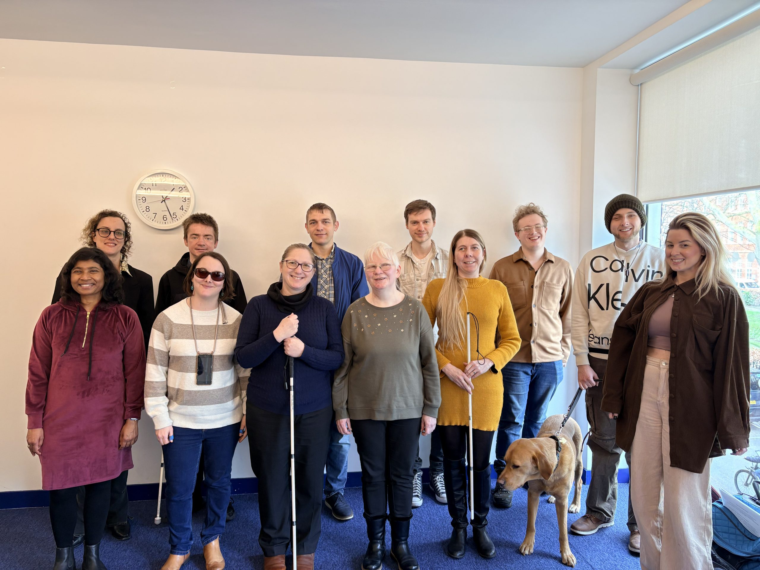 Group shot of 12 male and female interns from the 2025-26 cohort stood at the back of an office room with a blue carpet and cream walls. A guide dog can be seen in the photo. Several of the interns are holding a white navigation cane. They are smiling and look happy.