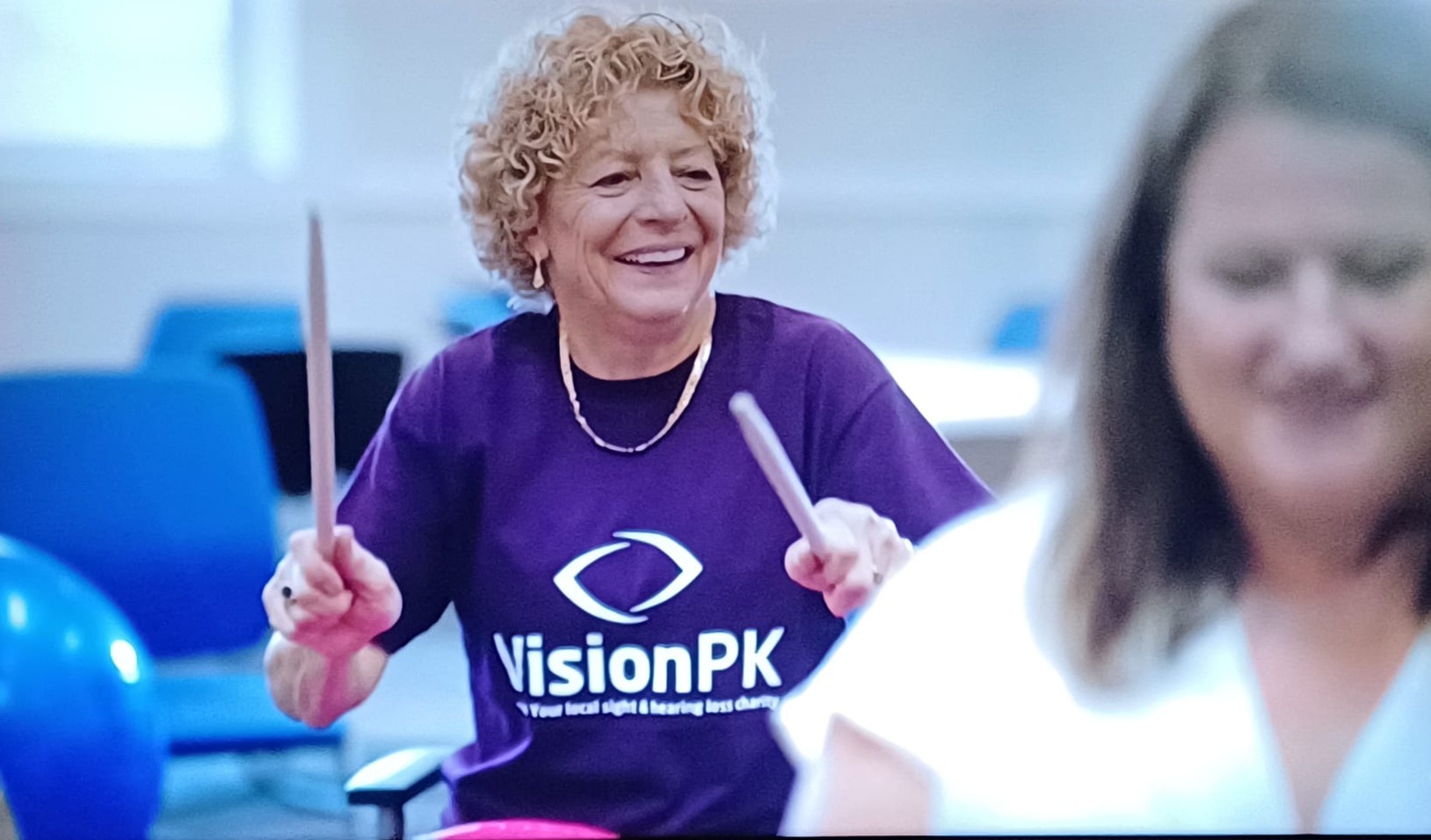 Two women smiling. The woman in the background is wearing a VisionPK T-shirt and holding up drumsticks.