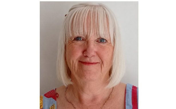 Andrea is smiling. She has a white bob with a fringe. She is wearing a gold, chain necklace and colourful top. 