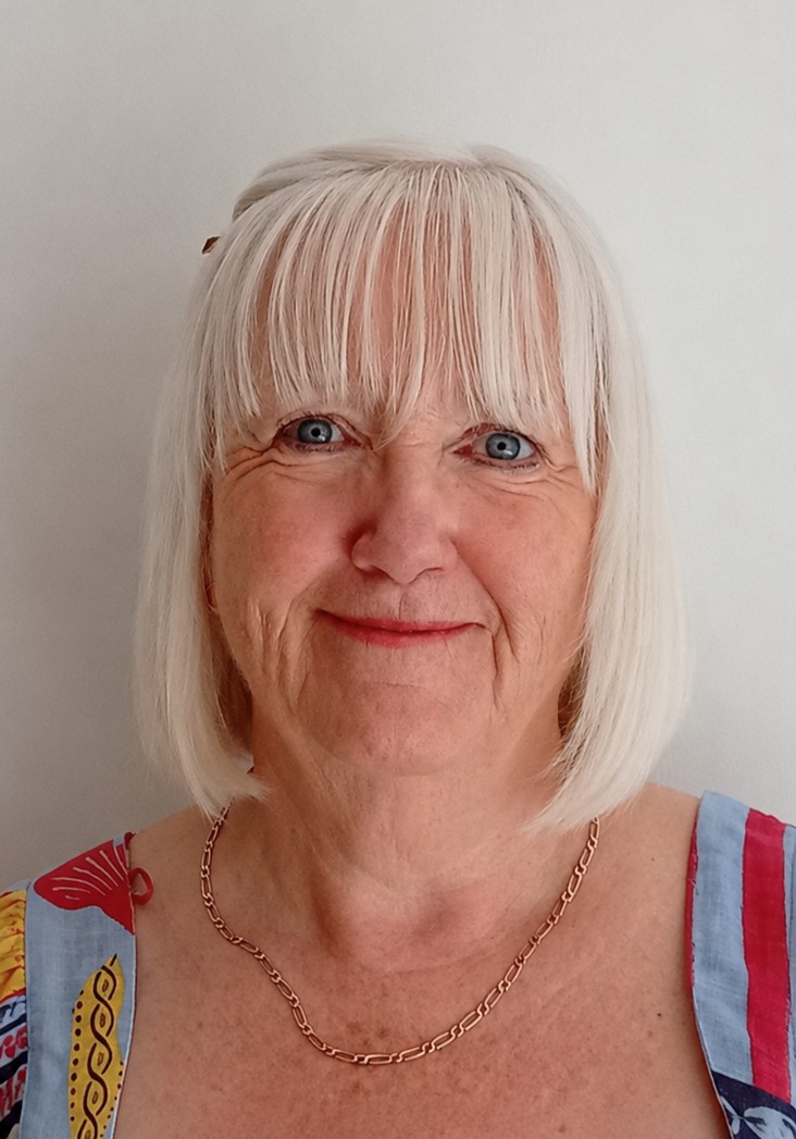 Andrea is smiling. She has a white bob with a fringe. She is wearing a gold, chain necklace and colourful top.