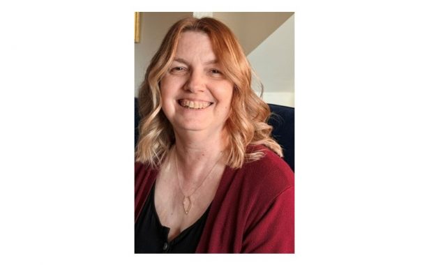 Barbara has a warm smile. She has shoulder length wavy auburn hair and is wearing a gold diamond shaped necklace, a black top and maroon cardigan.