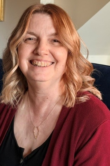 Barbara has a warm smile. She has shoulder length wavy auburn hair and is wearing a gold diamond shaped necklace, a black top and maroon cardigan.