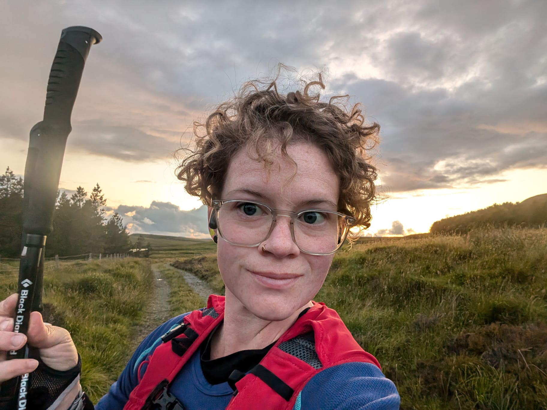 Photo by Wignall family. Shows Bri Wignall a blue top and carrying a red rucksack and a black walking pole, training for her ultramarathon on a hill track, with the sun setting behind her.