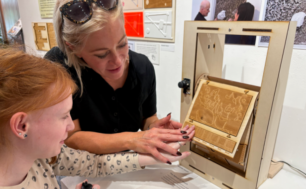 A young girl feels a gold, tactile artwork with the assistance of a woman.