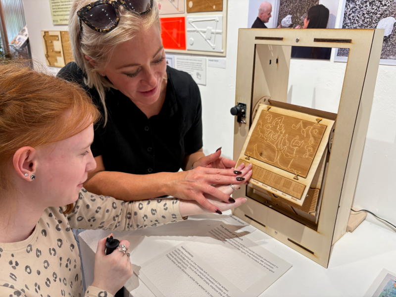 A young girl feels a gold, tactile artwork with the assistance of a woman.