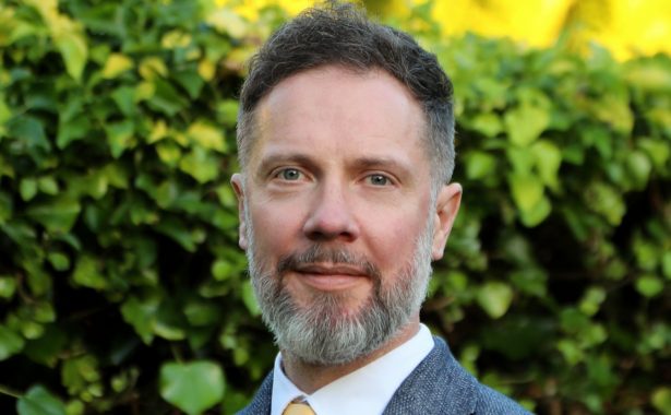 Sandy is smiling, standing outdoors with shrubs in the background. He has salt and pepper beard and moustache and is wearing a grey blazer, white shirt with a yellow tie.