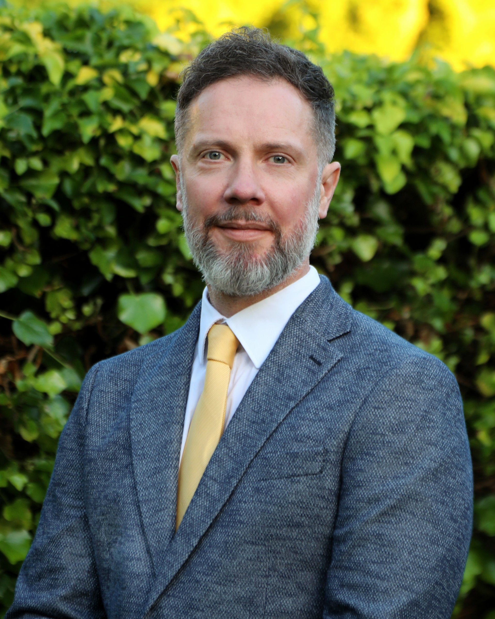 Sandy is smiling, standing outdoors with shrubs in the background. He has salt and pepper beard and moustache and is wearing a grey blazer, white shirt with a yellow tie.