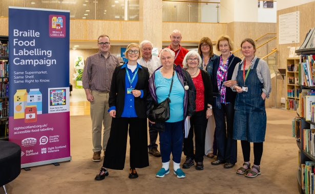 A group of nine people stand smiling together in the bright, open foyer of the Mitchell Library, with bookshelves on either side and a mezzanine balcony behind them. To the left is a tall purple and pink pull-up banner reading "Braille Food Labelling Campaign" with the slogan "Same Supermarket. Same Shelf. Same Right to Know." It shows illustrations of a juice carton, a milk carton and a jam jar, each marked with braille dots, alongside a scannable code, the web address for the campaign page, and the logos of Oban and District Access Panel, Disability Equality Scotland, Sight Scotland and Sight Scotland Veterans. The group is a mix of ages, dressed casually, with several people wearing lanyards.