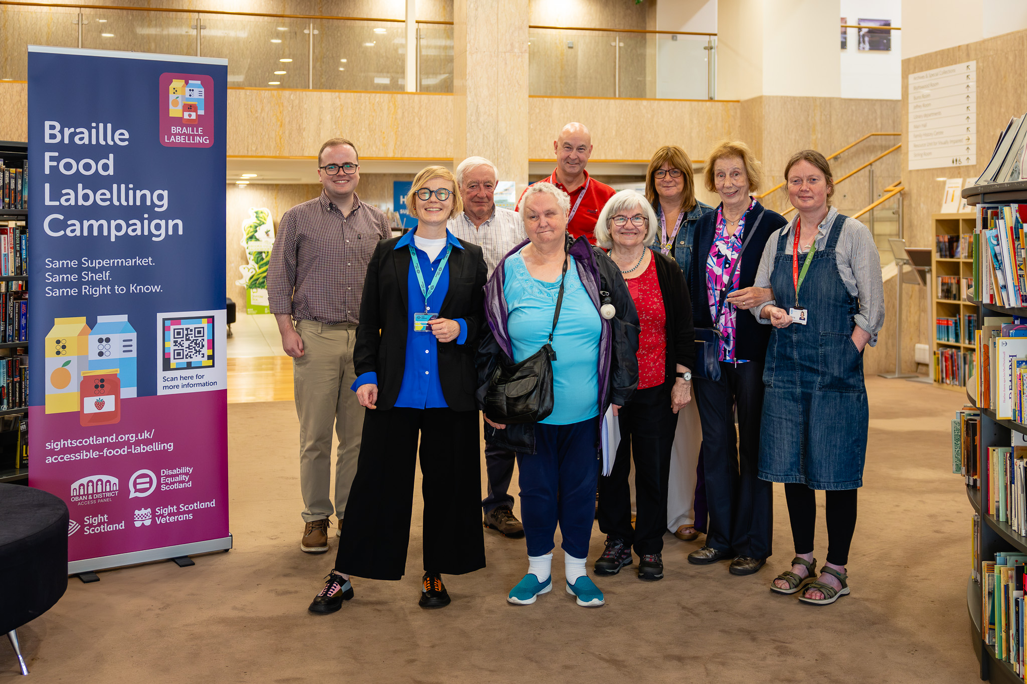 A group of nine people stand smiling together in the bright, open foyer of the Mitchell Library, with bookshelves on either side and a mezzanine balcony behind them. To the left is a tall purple and pink pull-up banner reading "Braille Food Labelling Campaign" with the slogan "Same Supermarket. Same Shelf. Same Right to Know." It shows illustrations of a juice carton, a milk carton and a jam jar, each marked with braille dots, alongside a scannable code, the web address for the campaign page, and the logos of Oban and District Access Panel, Disability Equality Scotland, Sight Scotland and Sight Scotland Veterans. The group is a mix of ages, dressed casually, with several people wearing lanyards.