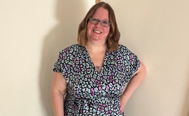 Gemma McKnight is smiling, standing with her left hand on her hip. She is wearing glasses and a silver/grey patterned dress.