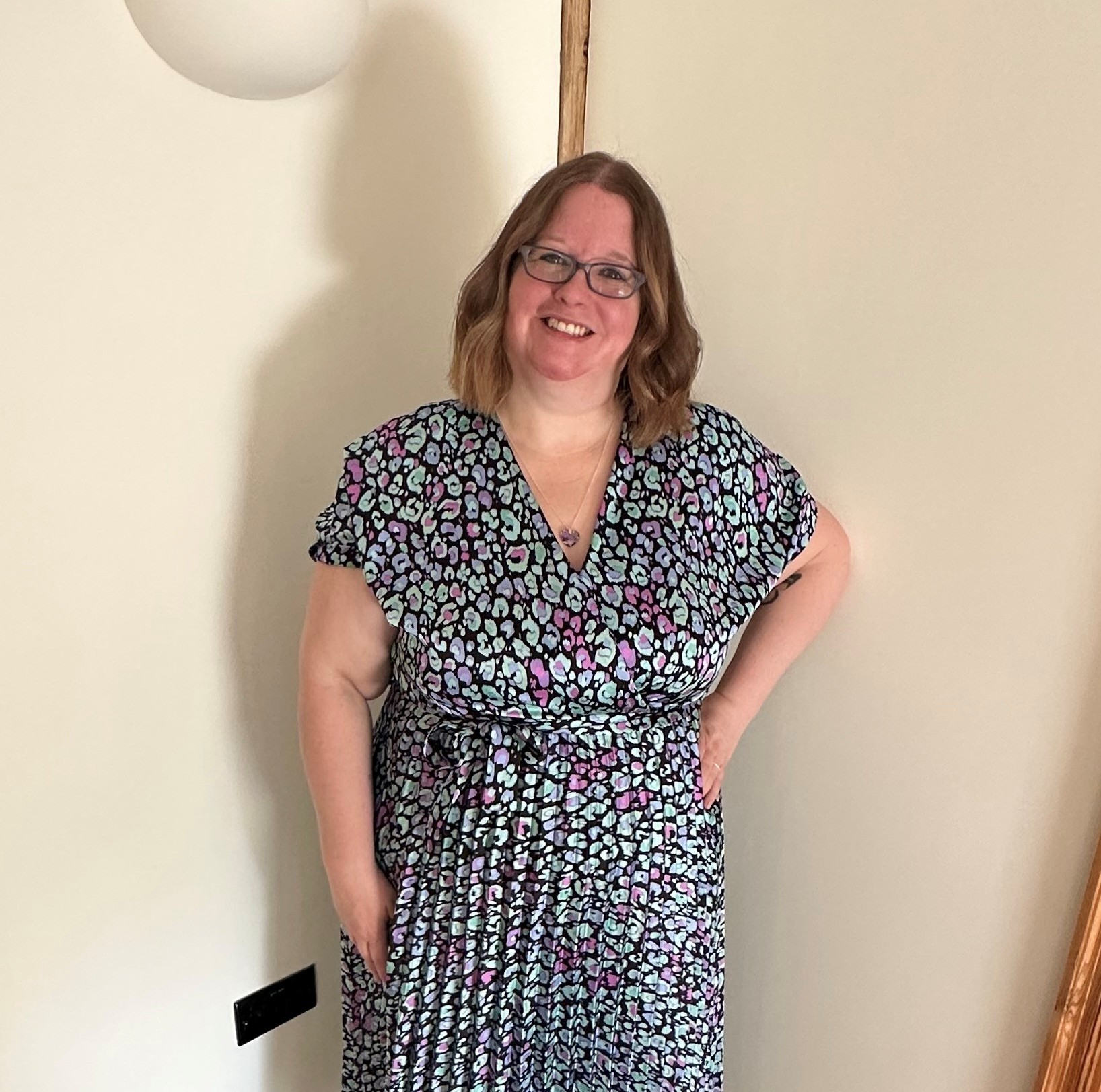Gemma McKnight is smiling, standing with her left hand on her hip. She is wearing glasses and a silver/grey patterned dress.
