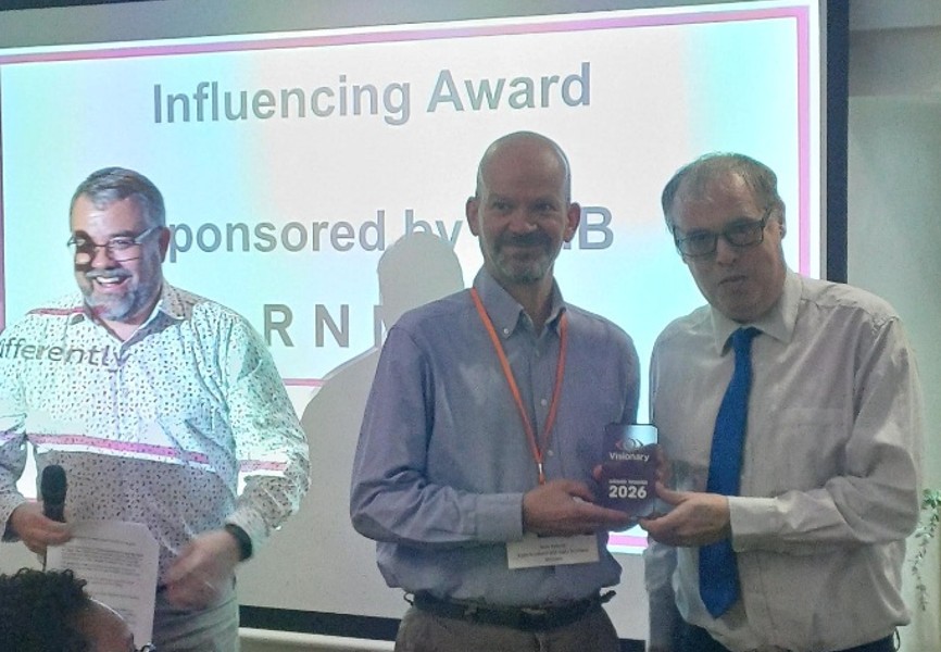 Craig Spalding from Sight Scotland and Sight Scotland Veterans is standing behind a lecturn. Mark Ballard from Sight Scotland Veterans is holding a trophy being presented to him by Mike Wordingham from RNIB.